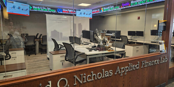 A view through glass into the David O. Nicholas Applied Finance Lab, showing desks, chairs, computer monitors, and digital stock ticker displays along the top wall.