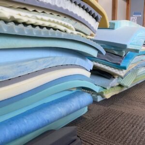 A stack of various foam mattress toppers and pads in different colors and textures is arranged on a carpeted floor near a wooden wall.
