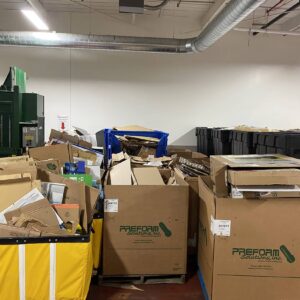 Large boxes and bins filled with flattened cardboard are arranged in a warehouse near a green cardboard baler and stacks of black containers.