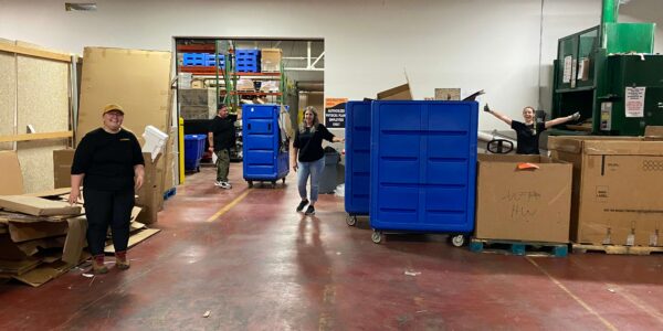Four people stand and pose in a warehouse among large blue carts and cardboard boxes on a red floor, surrounded by shelving and recycling equipment.