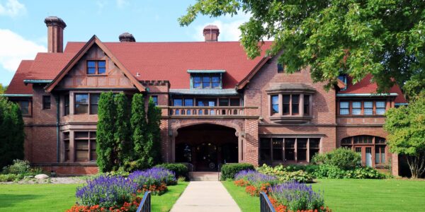 Large historic brick mansion with a red roof, arched entryway, manicured lawn, vibrant flower beds, and trees surrounding the building.