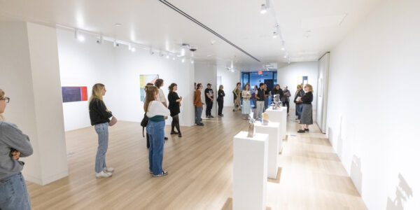 People stand and observe art pieces in a brightly lit modern gallery with wooden floors and white walls.