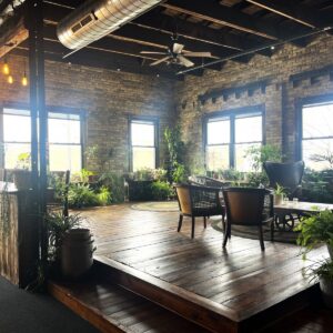 Industrial-style lounge with exposed brick walls, wooden floors, large windows, potted plants, a bar counter, and mixed seating arrangements under ceiling lights.