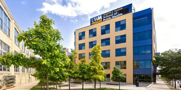 Modern four-story building with “UWM Joseph J. Zilber School of Public Health” sign, surrounded by young trees and a sidewalk under a partly cloudy sky.