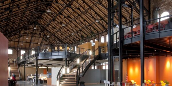 Spacious industrial-style office with exposed beams, a mezzanine, large windows, orange walls, open seating, desks, and a curved staircase.