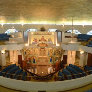 Interior of a circular church with blue pews, stained glass windows, and an ornate altar featuring religious icons and paintings under a domed ceiling.