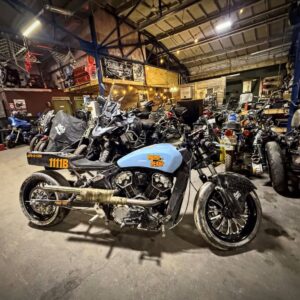 A custom blue motorcycle is parked in a cluttered workshop filled with various other motorcycles and garage equipment under bright ceiling lights.
