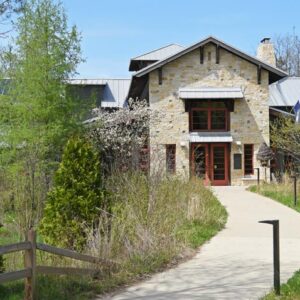 A stone and wood building with a metal roof is surrounded by greenery and trees, with a concrete path leading to the entrance.