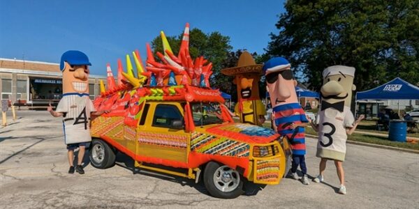 A small yellow truck decorated with traffic cones and bright designs is parked, surrounded by five people in oversized sausage costumes.
