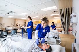 A nurse in blue scrubs checks a patient’s IV in a hospital room, while two other healthcare professionals work in the background.
