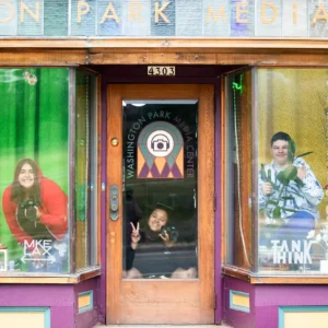 Three people pose inside a storefront window and door labeled “Washington Park Media Center.” The two windows display plants, decorations, and text: “MKE” on the left, “TANI” on the right.