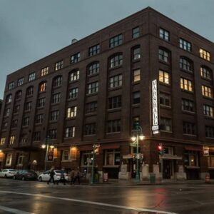 A large brick building at a street corner during dusk, with several windows lit and a vertical sign reading “MARSHALL” on the facade. Cars and pedestrians are visible nearby.