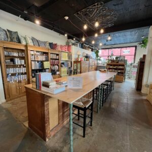 A spacious room with a long wooden table, bookshelves filled with books and pillows, bar stools, and large windows letting in natural light.