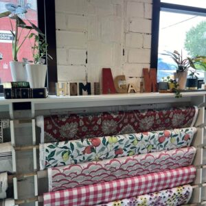 Fabric rolls with floral and plaid patterns are displayed on a white rack; letter blocks above spell “HOME AGAIN” beside plants and books.