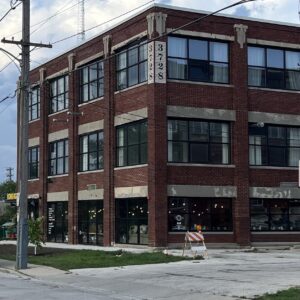 Three-story red brick building on a street corner with large windows, a street sign reading “3728,” and a construction barrier on the sidewalk.
