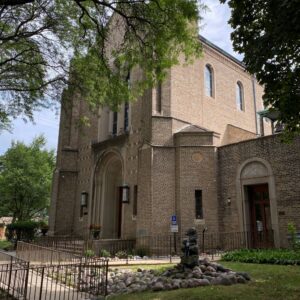 A brick church building with arched windows and doorways, surrounded by trees, a rock garden, and a wheelchair accessible ramp.