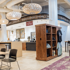 Spacious modern office lounge with tables, chairs, sofas, large hanging lights, a wooden bookshelf, and a visible kitchen area in the background.