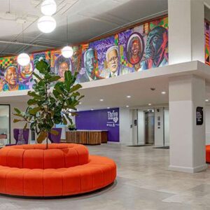 Modern lobby with orange circular seating, a large plant, purple chairs, and a colorful mural of faces on the upper wall. Stairs and reception area are visible in the background.