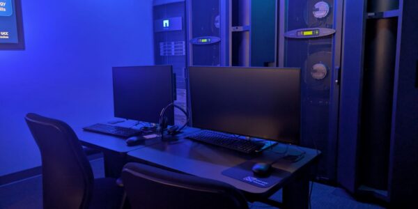 Two black office chairs and desks with monitors, keyboards, and headsets in front of tall server racks in a dimly lit, blue-toned room.