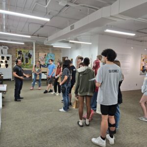 A group of people listens to a guide in an art gallery, surrounded by paintings on white walls and bright lighting from the ceiling.