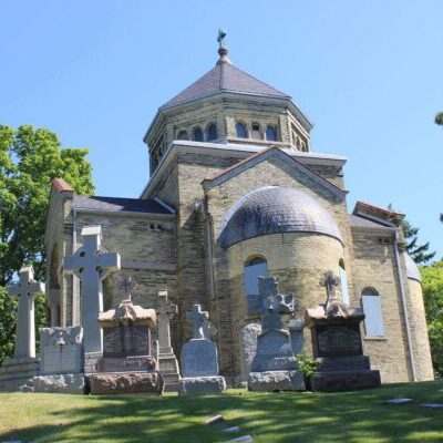 Stone mausoleum with a domed roof, surrounded by several large grave markers and trees, on a sunny day.