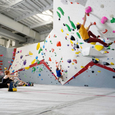 Three people watch as a climber ascends a colorful indoor bouldering wall in a brightly lit climbing gym.