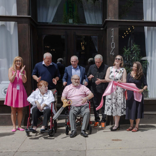 Eight adults gather outside a building for a ribbon-cutting ceremony; one person in a wheelchair holds oversized scissors cutting a pink ribbon.