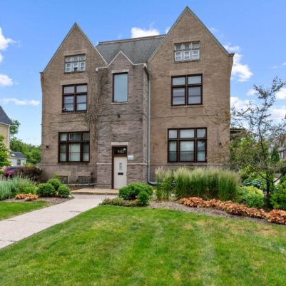 Two-story stone house with steep gables, large windows, and a central door, surrounded by landscaped lawn and shrubs on a clear day.