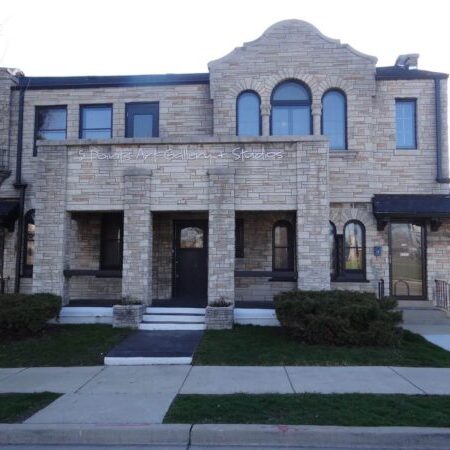Two-story stone building with arched windows and doorways, labeled “Spotlight Gallery Studios” above the entrance, fronted by shrubs and a sidewalk.