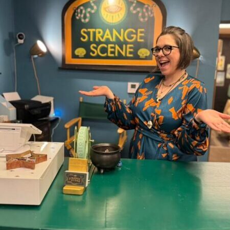 A woman in glasses and a patterned dress stands behind a green counter at a store called “Strange Scene,” smiling with arms raised in a welcoming gesture.
