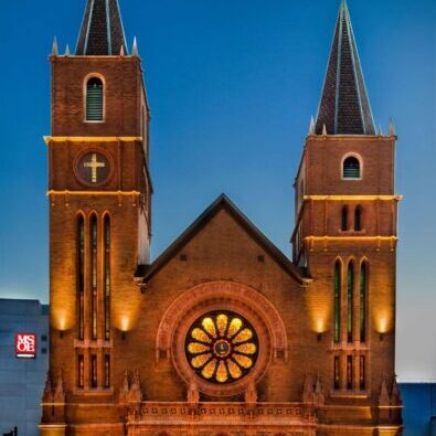 A large brick church with two tall steeples, arched doorways, and a circular stained glass window, lit up at dusk.