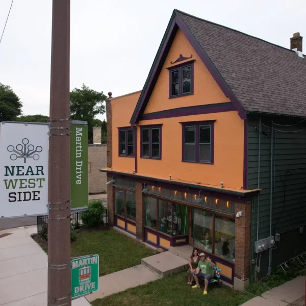 Two people sit on the front steps of a two-story orange and green house with large windows. A street sign reads “Near West Side Martin Drive.”.