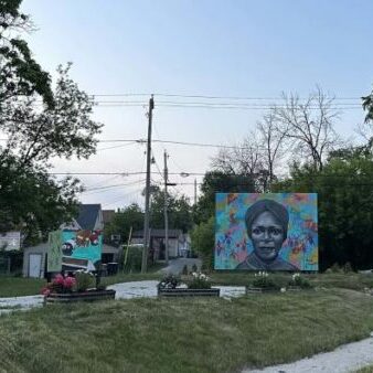Three colorful outdoor murals, including a portrait of a woman, are displayed on a grassy roundabout surrounded by trees and small houses under a clear sky.