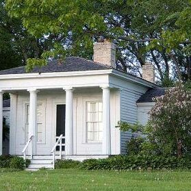 Small white house with four columns, black roof, and two chimneys, surrounded by green trees and shrubs on a grassy lawn.