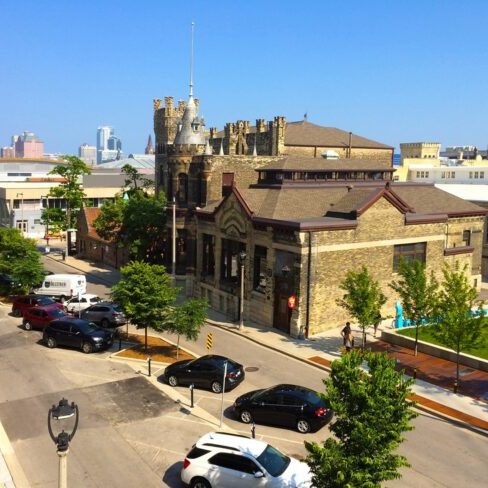 Street view of a historic stone building with castle-like features, parked cars, trees, and modern city buildings in the background under a clear blue sky.