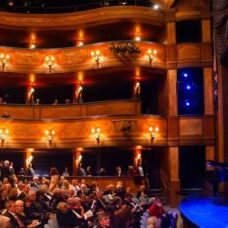 A full audience waits inside a historic theater with balcony seating and a curtain displaying the word “Skylight.” Musical instruments are set up on stage.