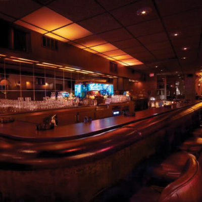 Dimly lit bar with leather stools, a polished wooden counter, mirrored wall, glassware, low lighting, and a TV displaying colorful visuals behind the bar.