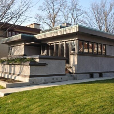 A low, gray modernist house with flat roofs, horizontal lines, and many windows stands on a green lawn with bare trees in the background.