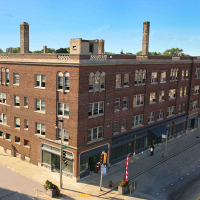 A four-story brick apartment building with retail spaces at street level, located on a corner with wide sidewalks and empty roads on a sunny day.