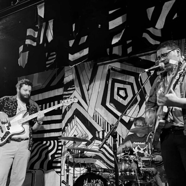 Three musicians perform on stage with guitars and drums in front of a bold, geometric black-and-white mural.