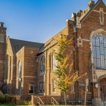 Large brick church building with tall windows, arched entrance, and a steep roof, surrounded by greenery under a clear blue sky.