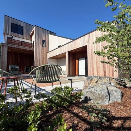 Modern two-story house with wood siding, large windows, and a patio featuring outdoor chairs and a table, surrounded by landscaping and clear blue sky.