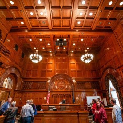 People stand and talk in a large, ornate wood-paneled courtroom with chandeliers, tall windows, and an American flag behind the judge’s bench.
