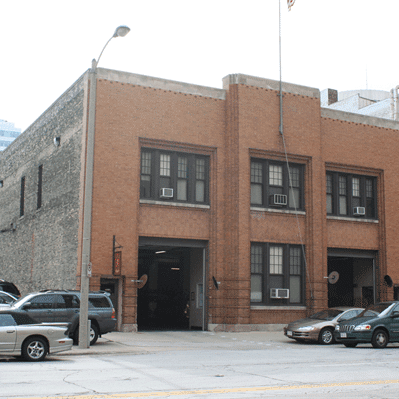 Two-story brick fire station with two garage doors, parked cars in front, and a Lowe’s billboard visible to the left.