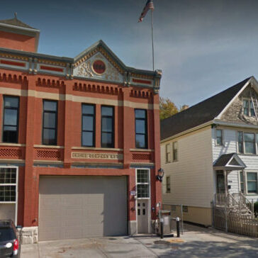 A two-story historic brick fire station with an arched garage door stands next to a white two-story house with a ladder on the side and a small fenced yard.