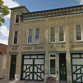 Two-story historic fire department building with tan brick, green-trimmed garage doors, and a small tower; a red and white house is adjacent on the left. The building is dated 1903.