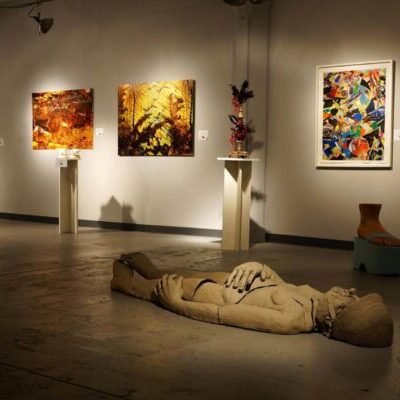 Art gallery with paintings on the wall, a yellow child statue, a reclining figure sculpture on the floor, and a large foot sculpture on display.