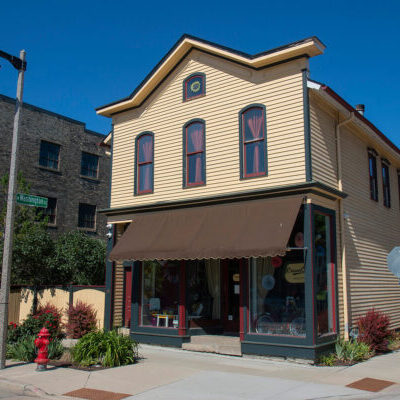 A two-story yellow building with brown trim and awnings sits on a street corner, surrounded by greenery and neighboring brick buildings under a clear blue sky.