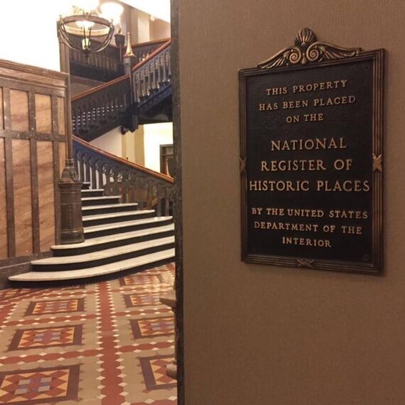 A plaque stating a property’s listing on the National Register of Historic Places is mounted on a wall next to a tiled floor and a staircase with ornate railings.