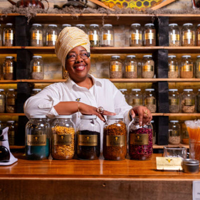 A woman stands behind a shop counter with large glass jars of herbs in front of her and shelves of smaller jars behind her.